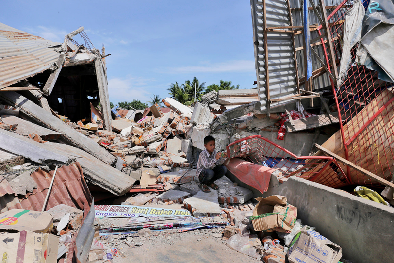 A boy sits on the rubble of a building that collapsed after an earthquake as he takes shelter from the sun in Pidie Jaya. (Image: AP/Heri Juanda)